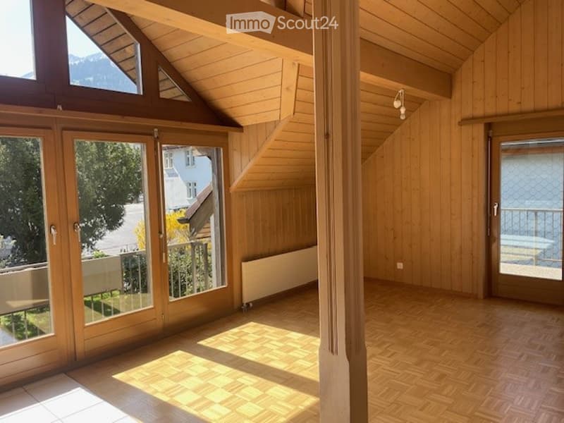 Bright, empty room with wooden floors and walls, large windows and sliding doors leading to a balcony, visible mountain view outside.