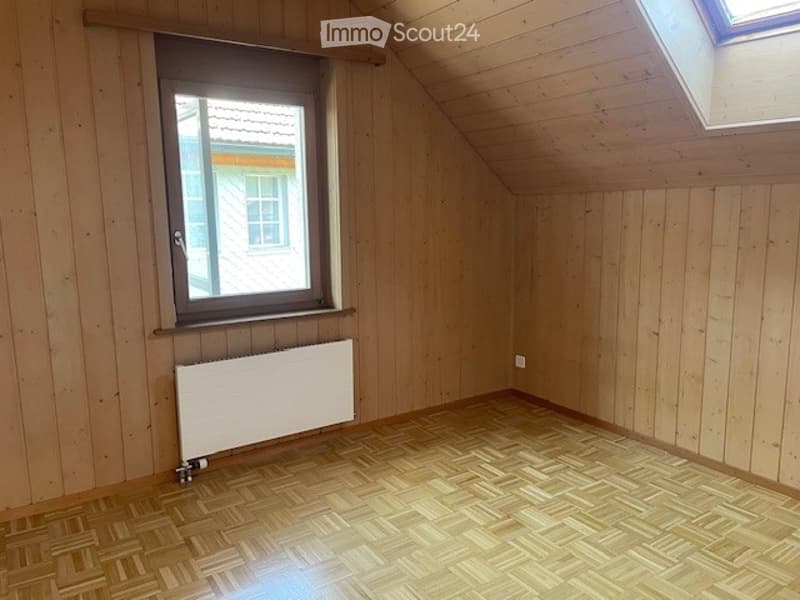 Empty attic room with wooden walls, herringbone wooden floor, large window, skylight, and radiator.