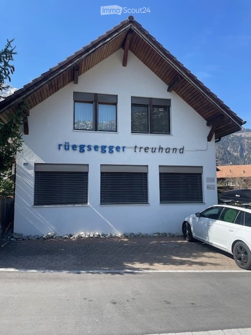 White building with a sloped wooden roof and multiple windows, featuring a sign that reads "rüggsegger treuhand". A white car is parked in front.
