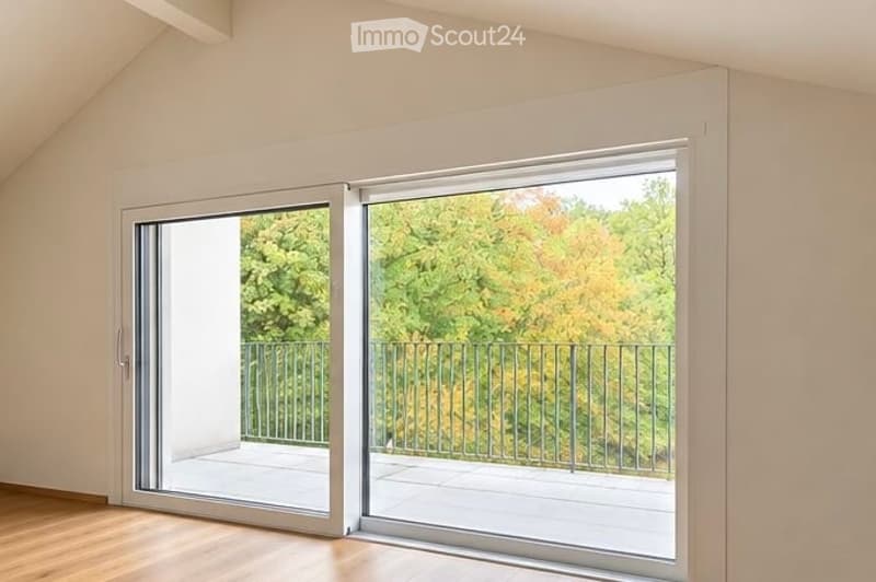 Bright, empty room with large sliding glass doors opening to a balcony, wooden floor, white walls, and a view of green trees.