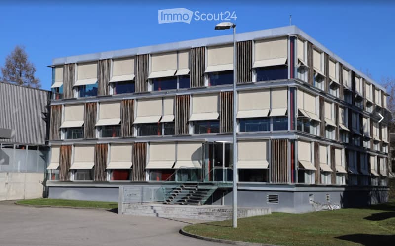 Modern multi-story building with a clean, geometric design, featuring large windows with roller shades and a raised entrance with steps.