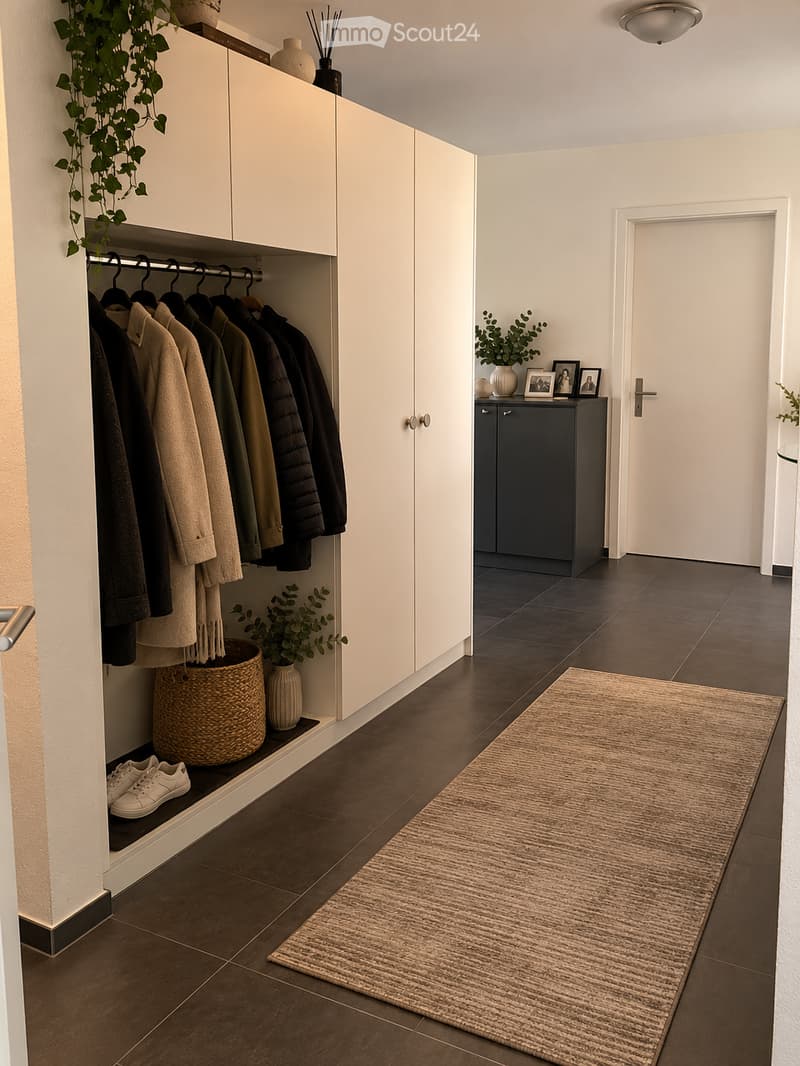 Modern hallway with dark tiled flooring, white built-in closet with hanging coats, small shelf with shoes and plants, a small black cabinet with framed photos, and a light-colored runner rug.