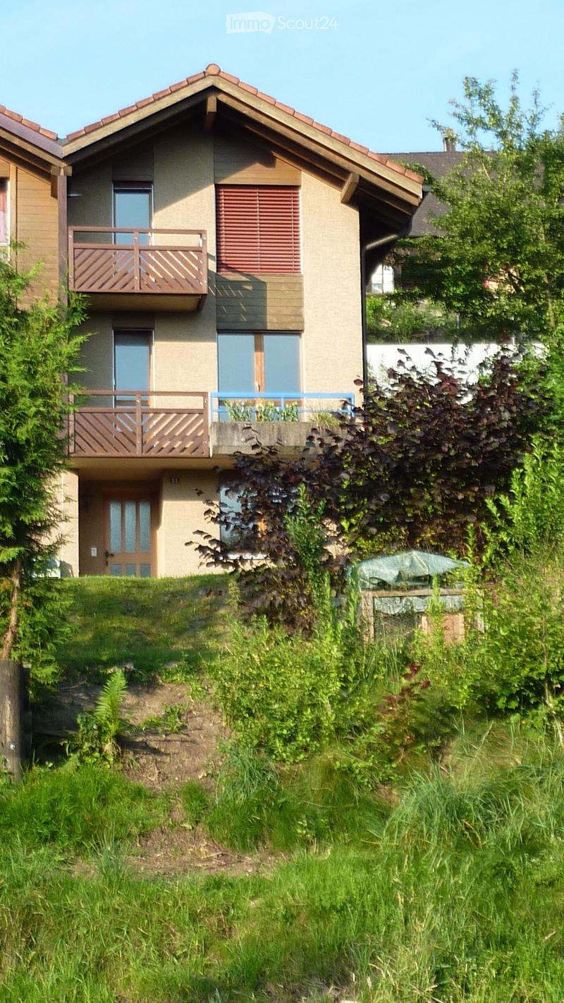 Two-story house with beige walls, wooden balconies, and multiple windows. The house is surrounded by greenery and has a raised ground floor.