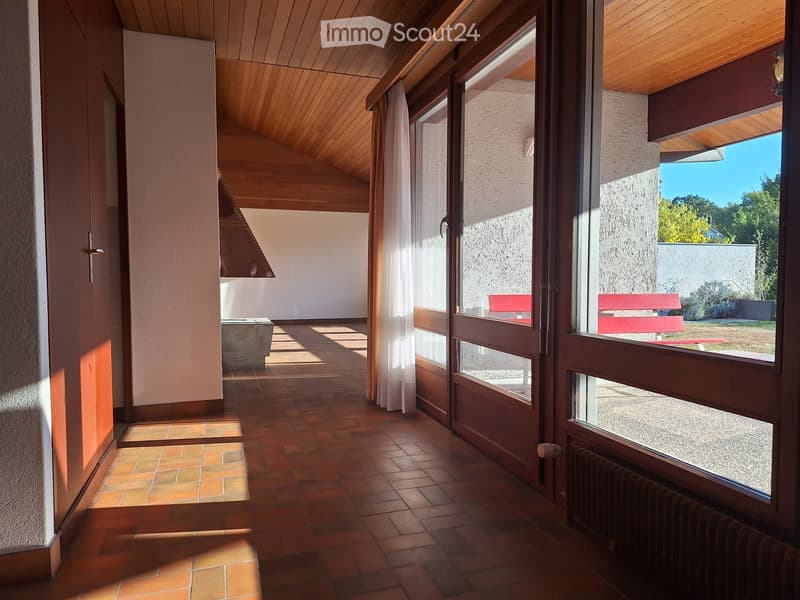 Bright hallway with wooden ceiling, tiled floor, large windows, and a view of a red bench outside.