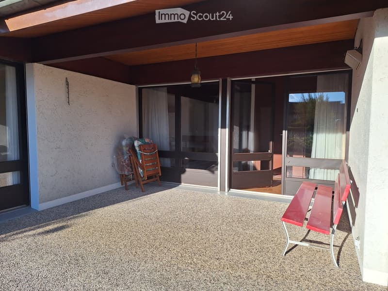 Outdoor patio with carpeted floor, wooden bench, and red chairs, with large sliding glass doors leading inside.