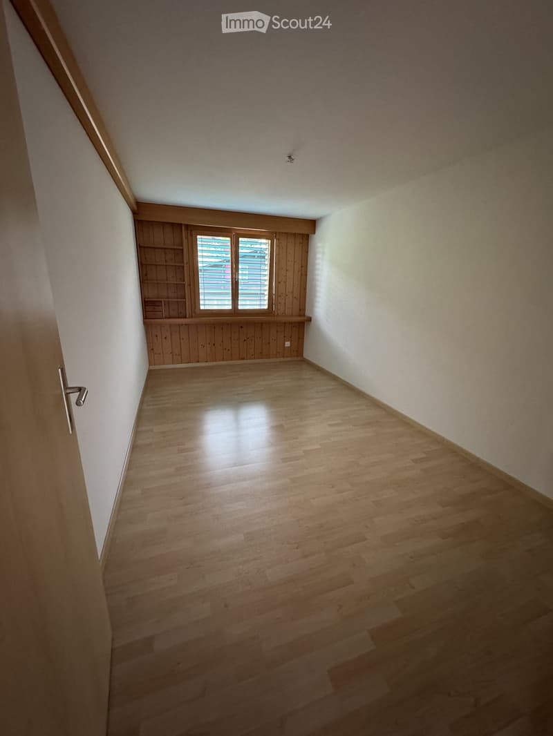 Empty room with wooden flooring, white walls, and a window with blinds. The room features built-in wooden shelving units and a door with a silver handle.