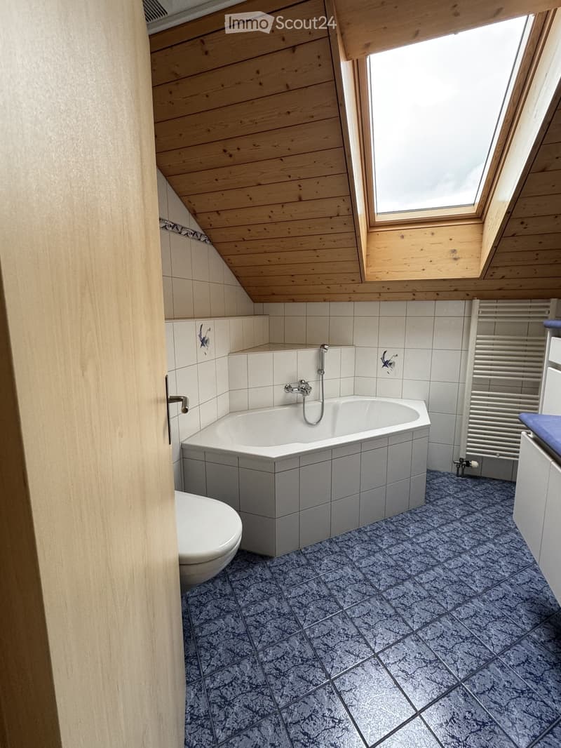 Bathroom with a corner bathtub, toilet, radiator, and skylight window. The walls are tiled, and the floor has a blue and grey patterned tile.