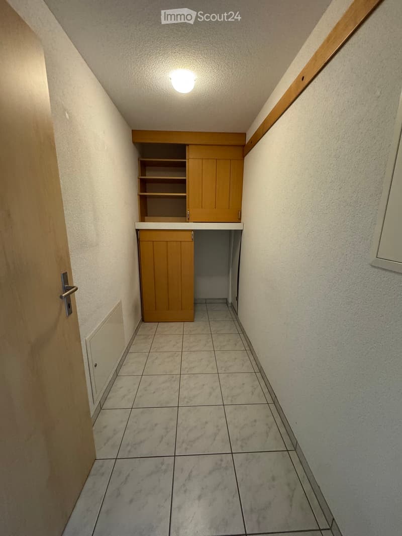 A narrow hallway with tiled flooring, light-colored walls, and a small wooden cabinet with shelves and a countertop on one side.