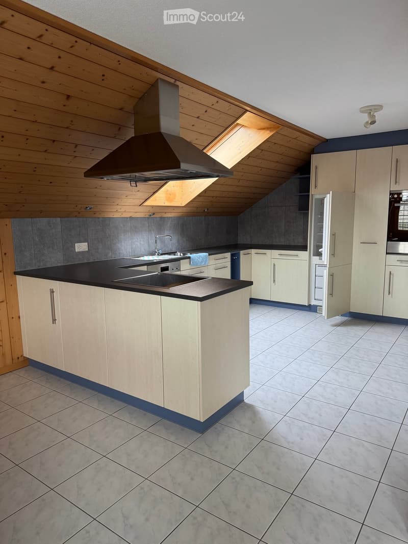 Modern kitchen with light wood cabinets, black countertops, a central island, and stainless steel appliances including a range hood, oven, and refrigerator. The space features a skylight and tiled flooring.