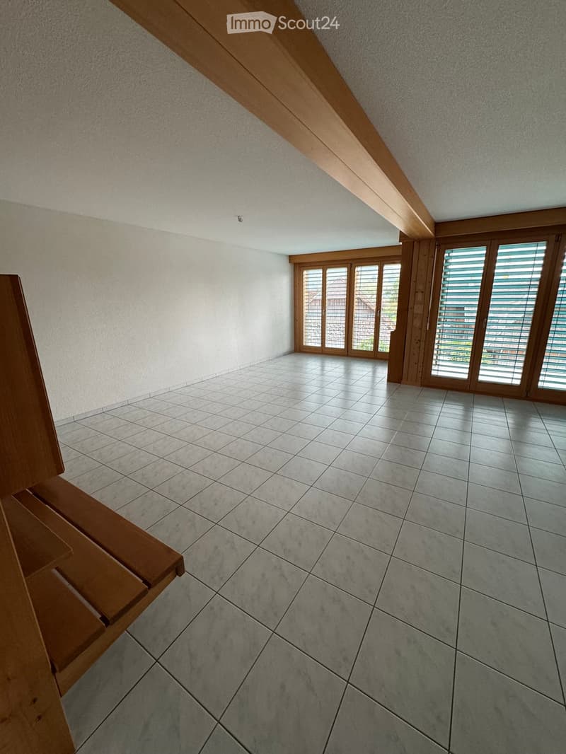Empty attic room with wooden beams, large windows, and tiled floor.