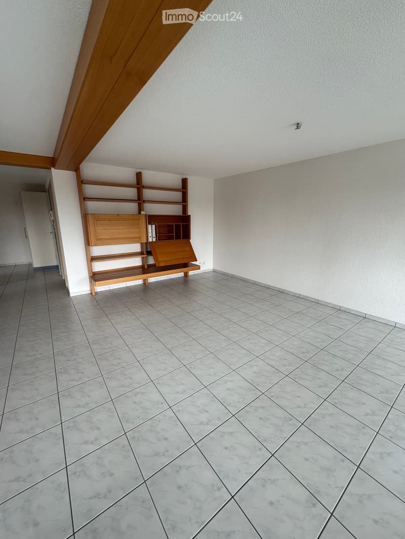 Empty room with light-colored walls, tiled floor, and a wooden shelf unit against one wall.