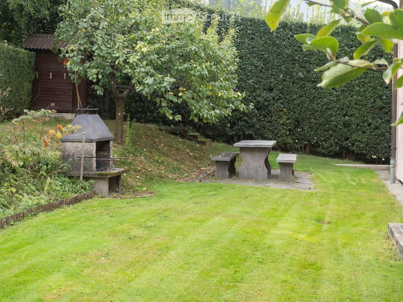 Well-maintained garden with a stone barbecue, stone benches, and a wooden shed, surrounded by lush greenery and hedges.