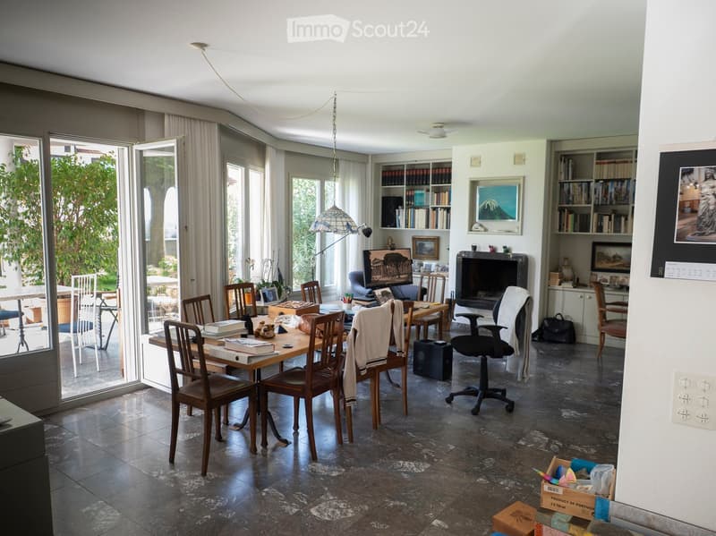 A spacious, well-lit room with a large glass door leading to a balcony, a wooden dining table with chairs, an office chair, a bookshelf, a fireplace, and a large screen.