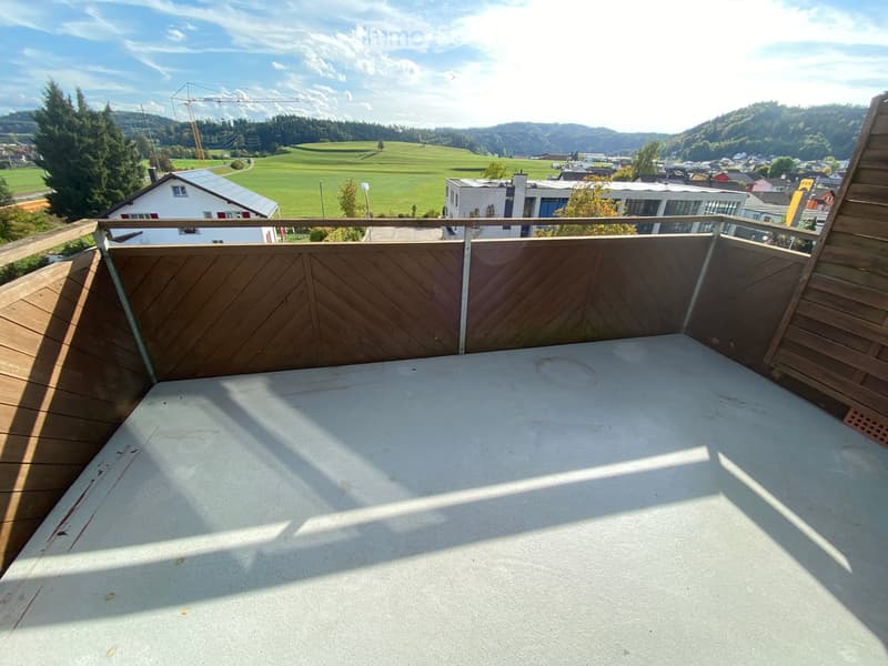 Empty wooden balcony with a metal railing, overlooking a green landscape and town with houses and trees.