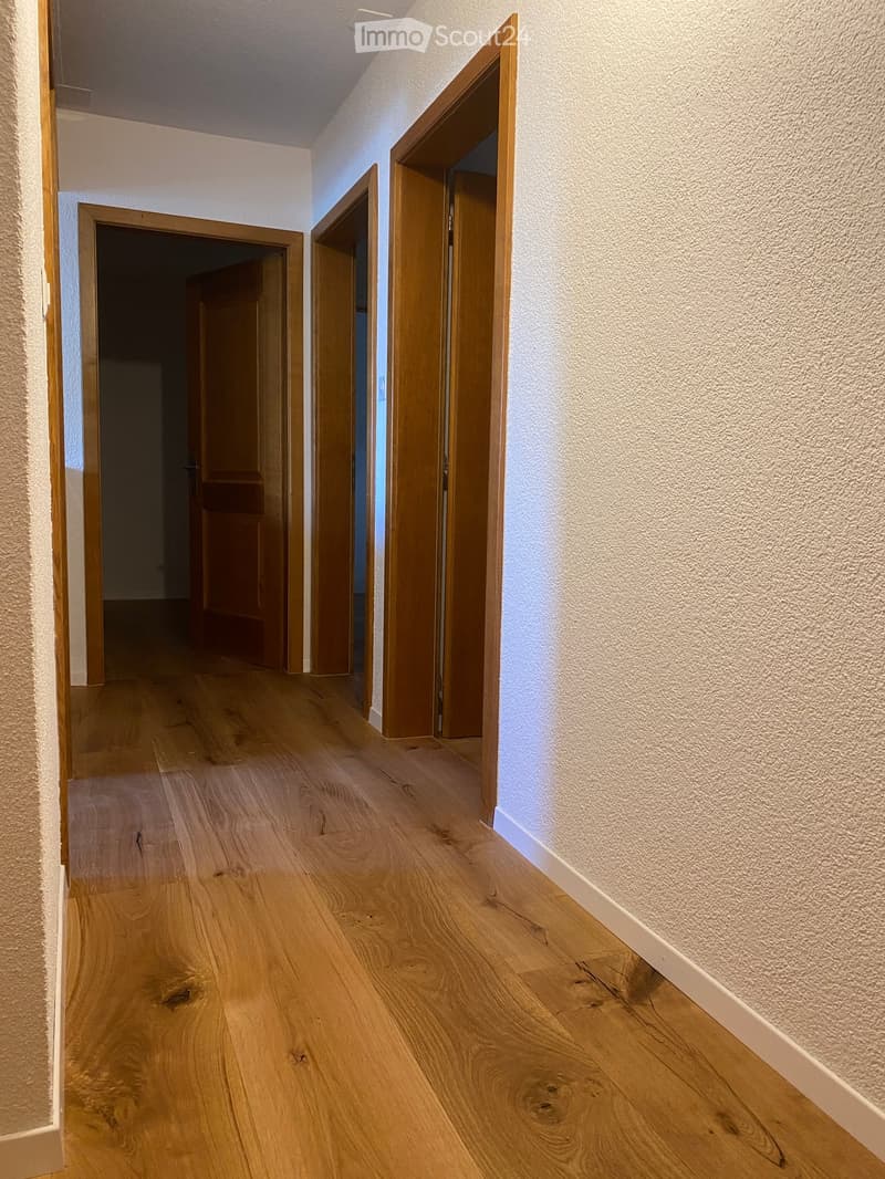 A clean, well-lit hallway with wooden flooring and white walls, featuring several open wooden doors leading to other rooms.