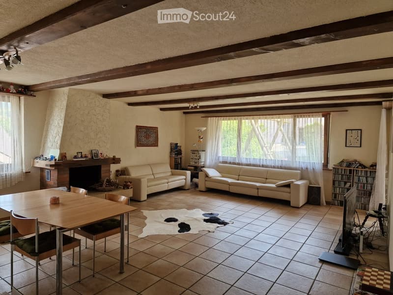 Living room with exposed wooden beams, tiled floor, two cream-colored sofas, a wooden dining table with four chairs, a fireplace with decorative items, a bookshelf, and a television setup.