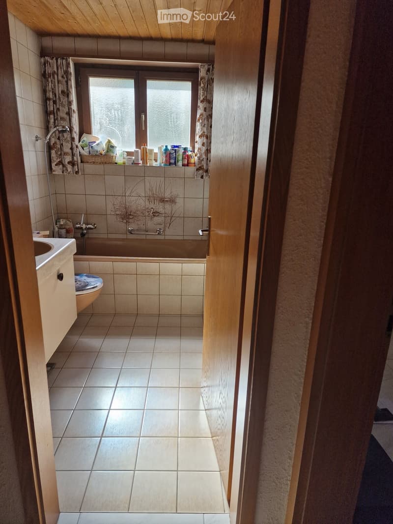 Bathroom with tiled walls and floor, a bathtub, toilet, sink, and a window with curtains. A basket with toiletries is placed on the windowsill.