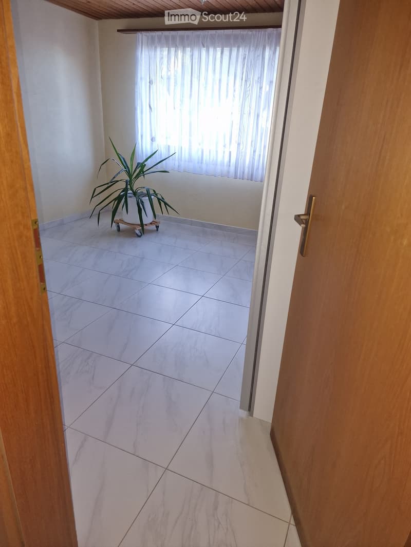 Empty room with white tiled flooring, white walls, and a window with sheer curtains. A small potted plant sits on a wheeled stand near the window.