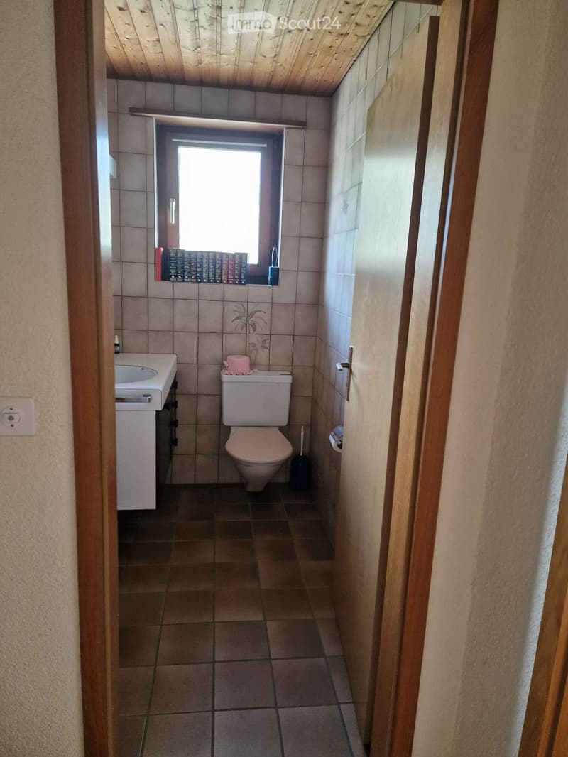 Small bathroom with tiled walls and floor, featuring a sink, toilet, and a window with books and a toothbrush on the sill.