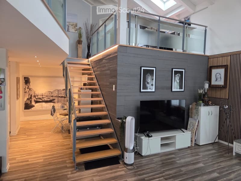 Modern living room with wooden flooring, staircase leading to an upper level, large flat-screen TV on a white stand, a white fan, and decorative picture frames on the wall.