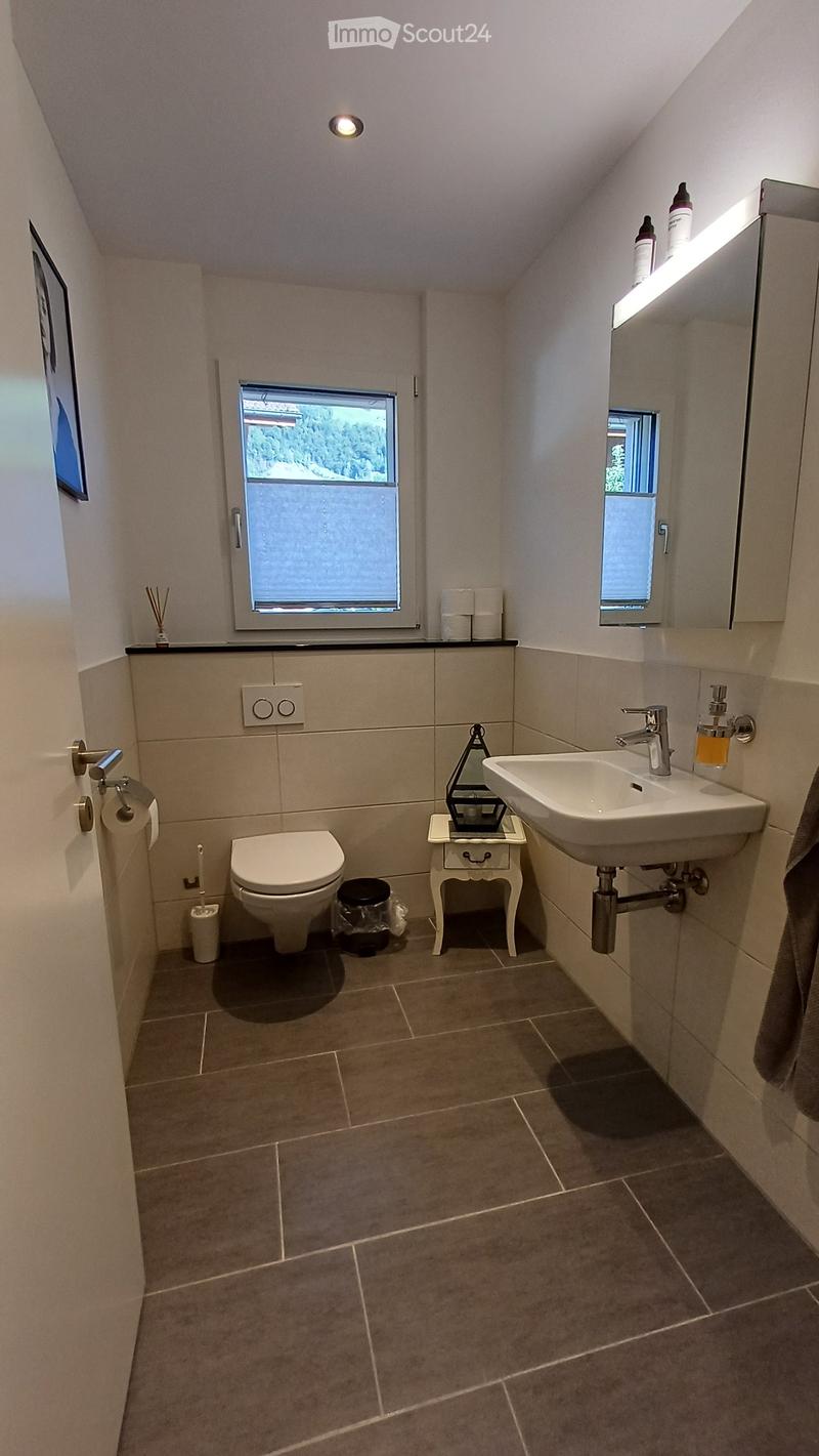 Modern bathroom with beige tiles, a toilet, a sink with a modern faucet, a small white stool, a mirror, and a window with frosted glass. The room features a sleek design with a built-in light fixture above the mirror.
