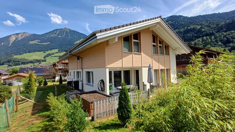 Two-story house with light brown siding and white trim, surrounded by greenery and overlooking a mountainous landscape.