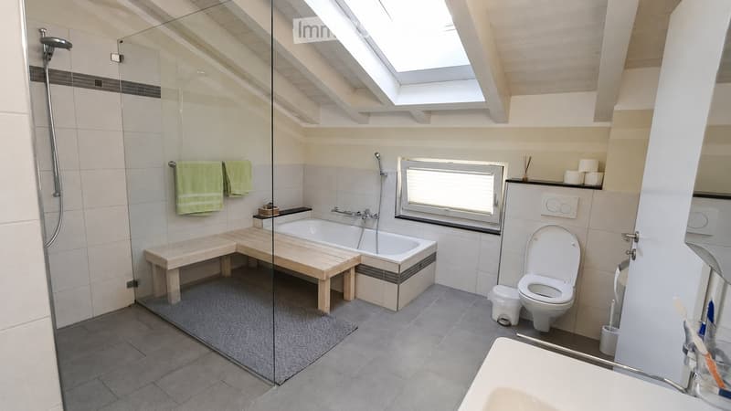 Modern bathroom with a bathtub, toilet, wooden bench, and glass-enclosed shower area. Features a skylight window and a small window, with towels and toiletries neatly arranged.
