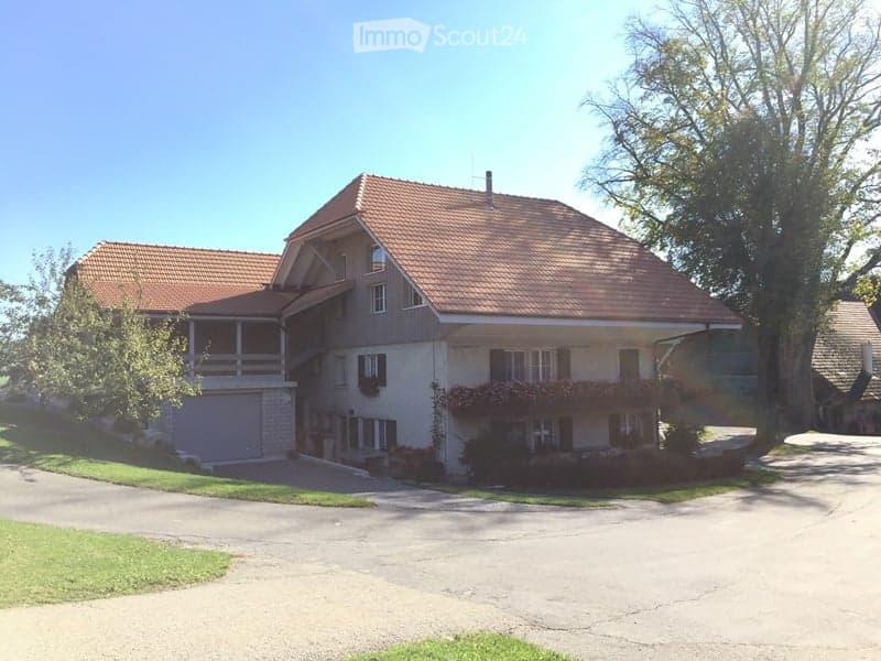 Two-story house with a red-tiled roof, white walls, and multiple windows. Features a covered balcony on the upper level and a garage on the left side.