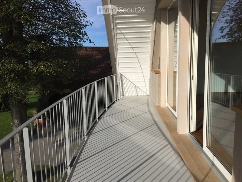 Balcony with white metal railing, wooden decking, large windows, and a view of a green lawn and trees.