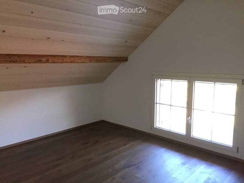 Empty room with wooden floor, white walls, and large windows allowing natural light to enter. The ceiling has exposed wooden beams.