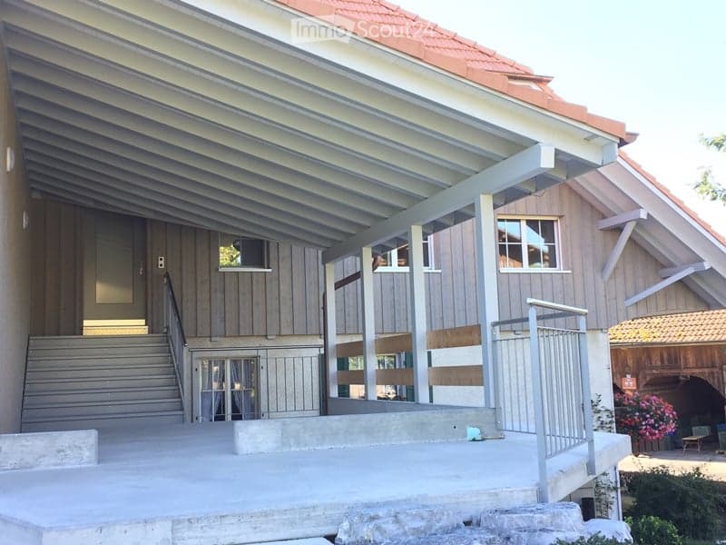 Modern building entrance with raised ground floor, wooden facade, metal railings, and steps leading up to the entrance.