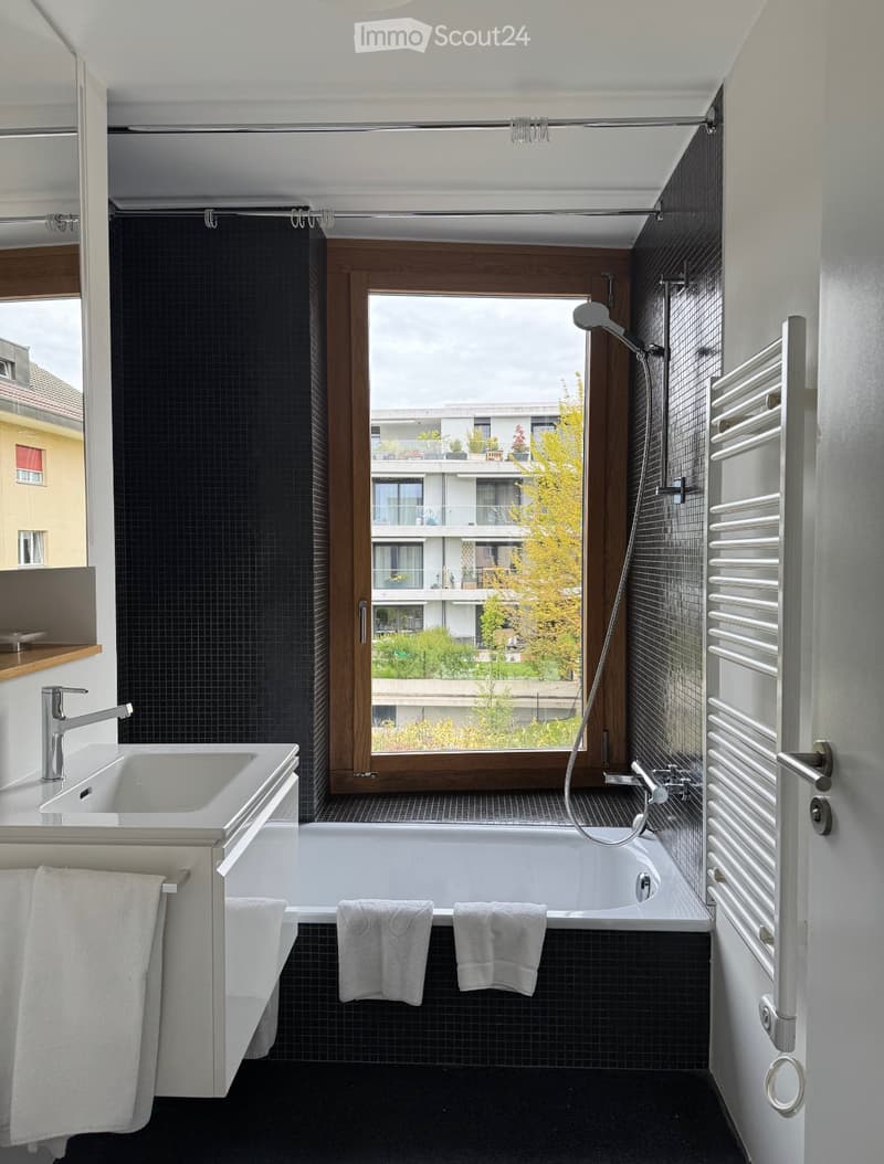 Modern bathroom with black tiled walls, a large window offering a view of the city, a white sink with a chrome faucet, a bathtub, and a towel rack with white towels.