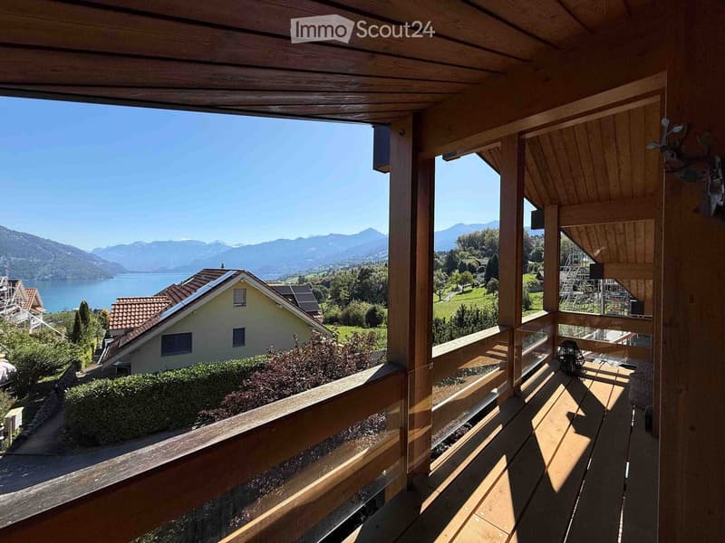 Wooden balcony with a railing, overlooking a scenic lake and mountain landscape.