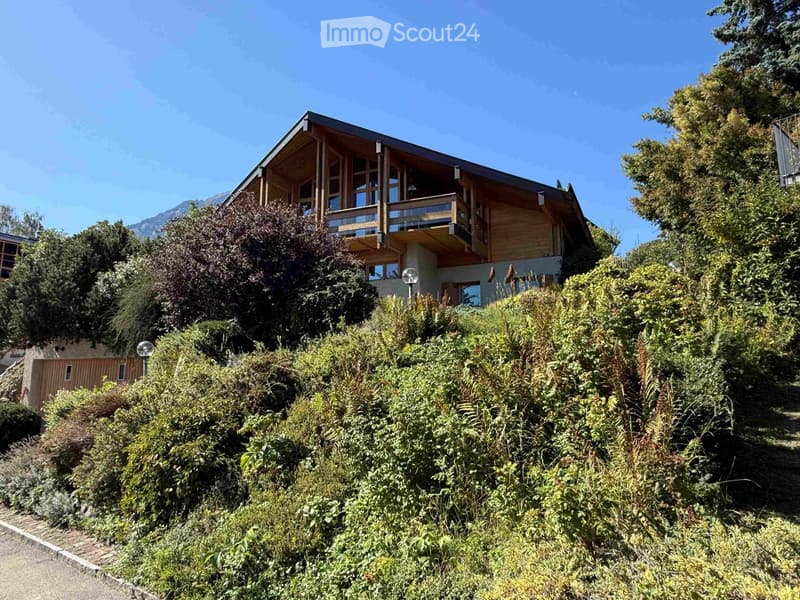 A two-story wooden house with large windows and a balcony, surrounded by lush greenery and trees.