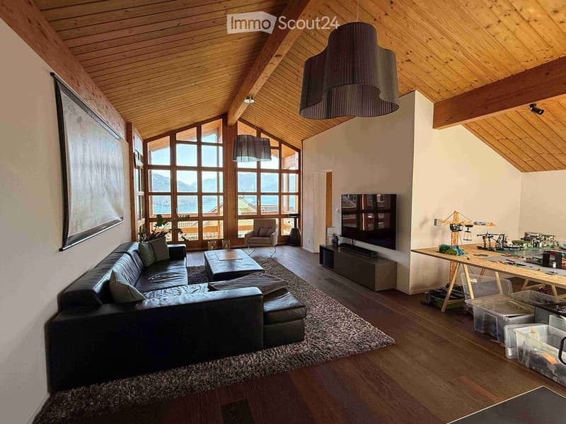 Modern living room with wooden ceiling, large windows offering a scenic view, sofa, coffee table, ottoman, chair, TV screen, and a table with toys.