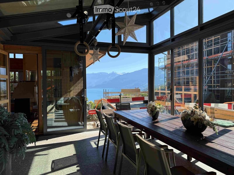 Bright, airy room with large windows offering a scenic mountain and lake view. Features a wooden dining table with chairs, flower vases, and a fireplace.