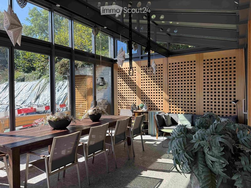 Bright, modern living space with large windows, wooden dining table, sofa, side table, and decorative plants.