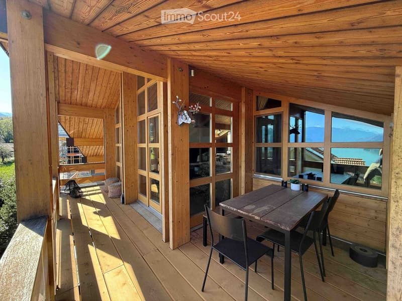 Wooden balcony with large windows offering a scenic view, featuring a table, chairs, a bench, and a small potted plant.