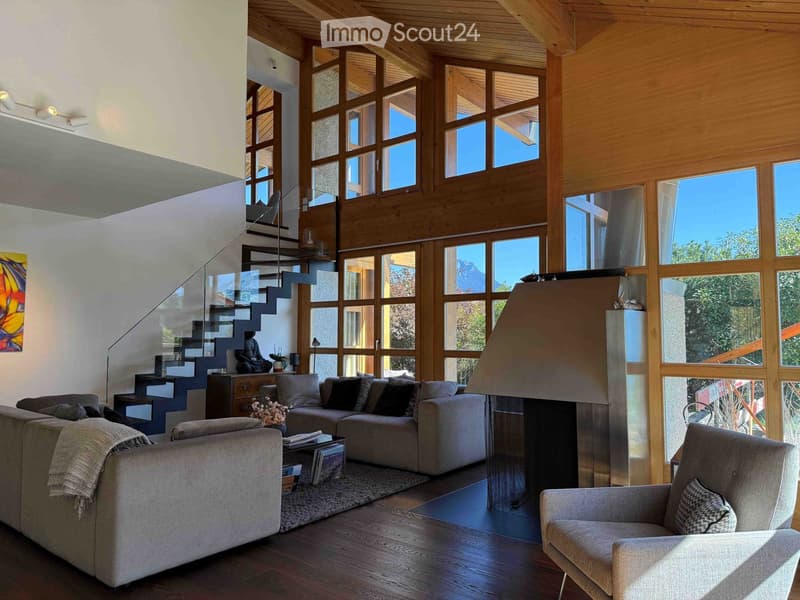 Modern living room with large windows, wooden ceiling, and a view. Features a grey sectional sofa, coffee table, armchair, fireplace, and a staircase leading to an upper level.