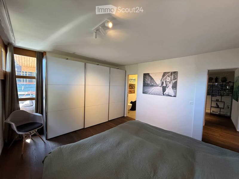 Modern bedroom with a large bed, a sleek white wardrobe, a contemporary chair, and a large framed picture on the wall. The room features wooden flooring and large windows with curtains.