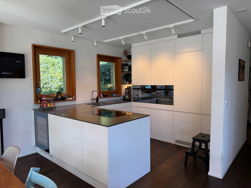 Modern kitchen with white cabinets, island, sink, and appliances. Features wooden flooring, large windows, and a small seating area with a table and chairs.