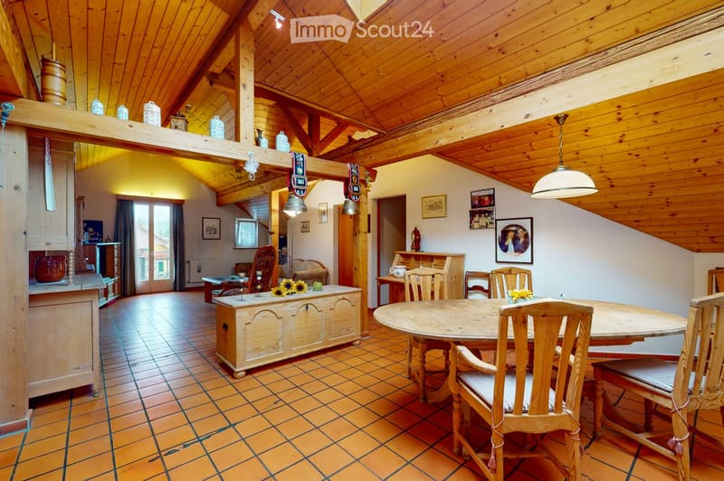 A cozy, wooden-beamed living and dining area with a round wooden dining table surrounded by chairs, a bench, and a small cabinet. The space features exposed wooden beams, warm lighting, and decorative items including vases and picture frames.