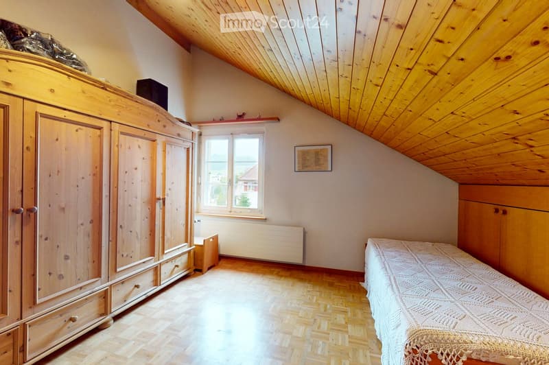 Cozy attic bedroom with wooden slanted ceilings, wooden floor, large window, bed with white bedspread, wardrobe, radiator, and framed picture on wall.