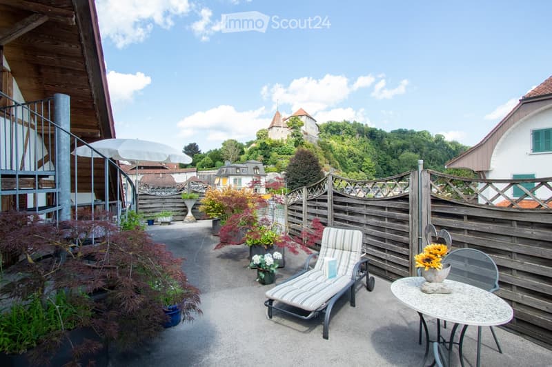 Balcony with a view of a town and surrounding greenery, featuring a lounge chair, small table, umbrella, and several potted plants.