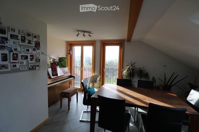 Bright dining area with a wooden dining table, chairs, a piano, and a small balcony with plants visible through the glass doors.