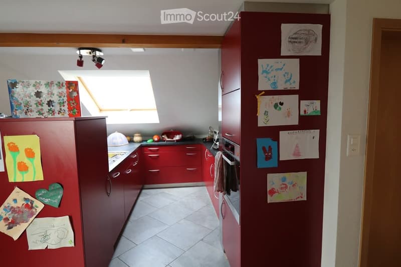 Modern kitchen with red cabinets, white tiled floor, and a skylight window. The refrigerator is decorated with children's drawings and artwork.