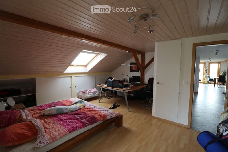 Spacious attic bedroom with wooden floors and slanted wooden ceilings. Features a large bed with colorful bedding, a desk with a computer and office chair, and built-in storage. Skylights provide natural light, and the room has a cozy, lived-in feel.