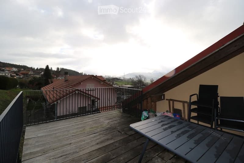 Wooden balcony with metal railing, overlooking a residential area with houses and mountains in the background. Includes a table and chairs.