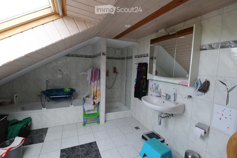 Bathroom with a bathtub, sink, mirror, shower, and various towels. The room features a slanted wooden ceiling and white tiled walls.