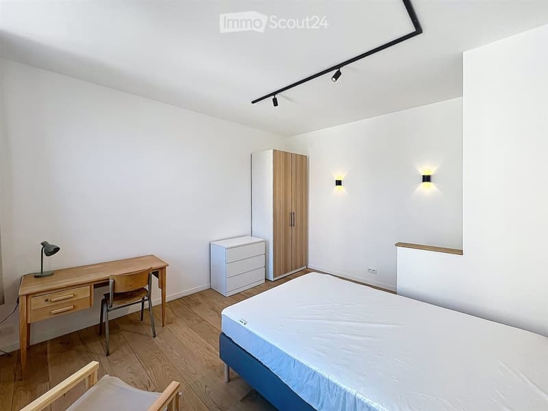 A tidy bedroom with a single bed, a wooden desk with a chair, a wardrobe, and a drawer unit. The room features modern lighting and a sleek design.